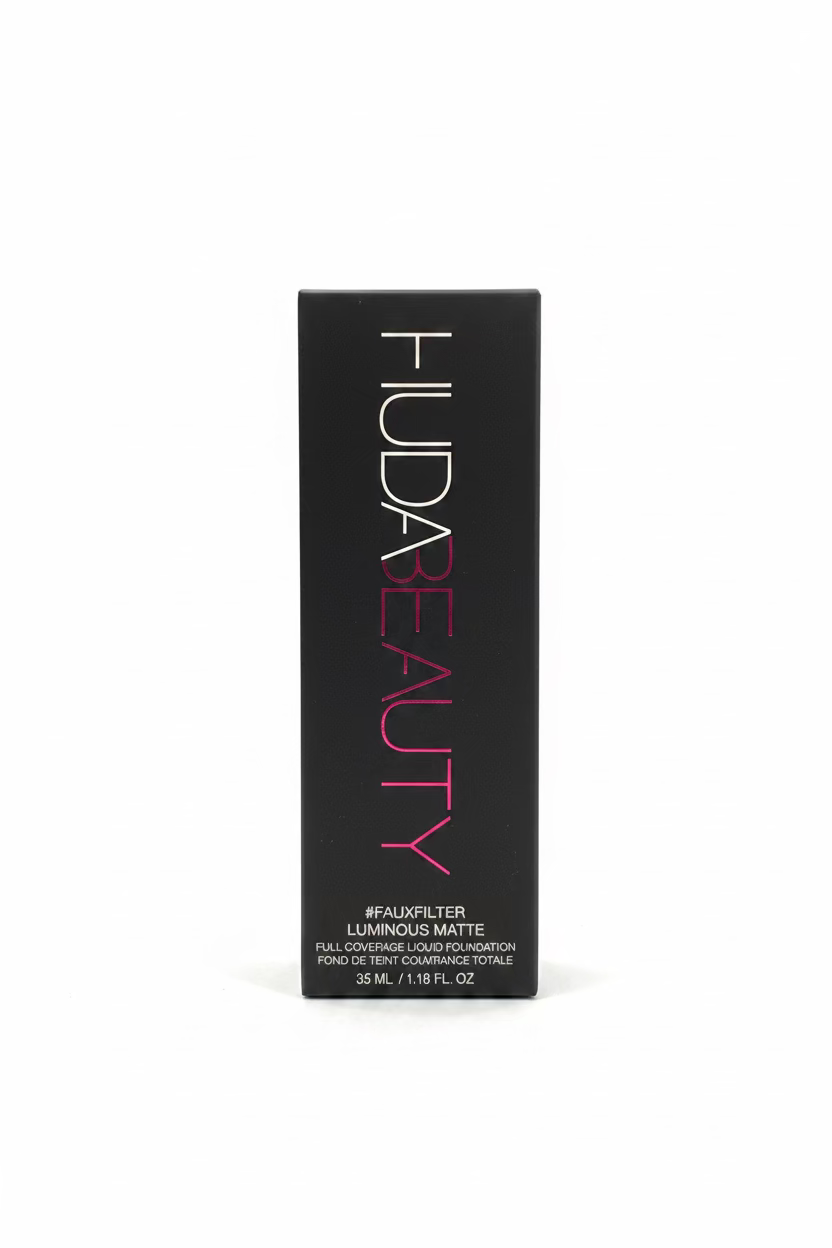 A black rectangular box for HUDA BEAUTY eyeshadow stands upright on a glass tabletop in a retail or storage environment. The box is labeled 'HUDA BEAUTY' with pink and white lettering, and the product name appears to be 'Luminous Matte'.