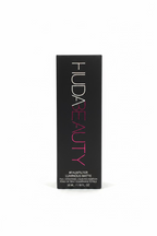 A black rectangular box for HUDA BEAUTY eyeshadow stands upright on a glass tabletop in a retail or storage environment. The box is labeled 'HUDA BEAUTY' with pink and white lettering, and the product name appears to be 'Luminous Matte'.