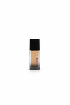 A bottle of Huda Beauty liquid foundation with a black cap and clear lower bottle showing beige foundation, standing on a glass counter in a busy retail setting with a laptop and Marvel boxes in the background.