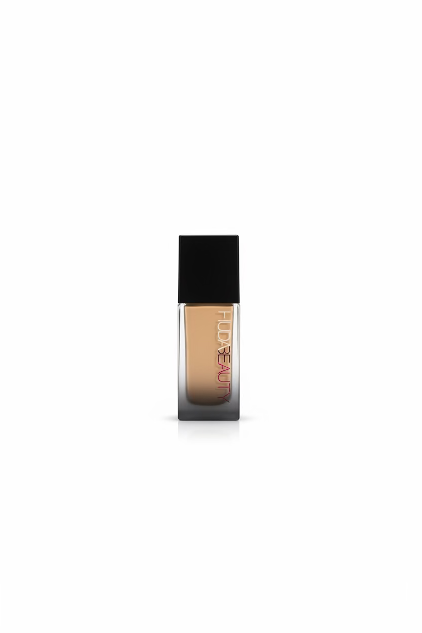 A bottle of Huda Beauty liquid foundation with a black cap and clear lower bottle showing beige foundation, standing on a glass counter in a busy retail setting with a laptop and Marvel boxes in the background.