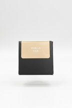 A hand holding a small black box with a beige label that reads 'VANILLA 120B' in a casual indoor setting.