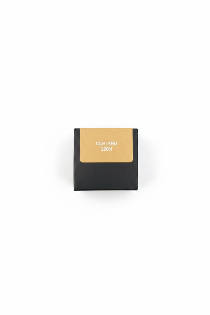 A hand holding a small product box with a tan color swatch labeled 'CUSTARD 220N' on the front, set against a background of gray denim jeans and a brown wooden surface.