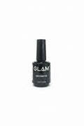 A hand holding a black bottle of GLAM Dehydrator nail polish against a background of blue jeans and a wooden cabinet.