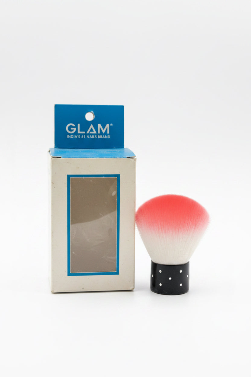 A small cosmetic brush with a red-to-white bristle gradient and a black polka-dot handle sits next to its blue and beige GLAM India nail brand box on a desk in front of a laptop.