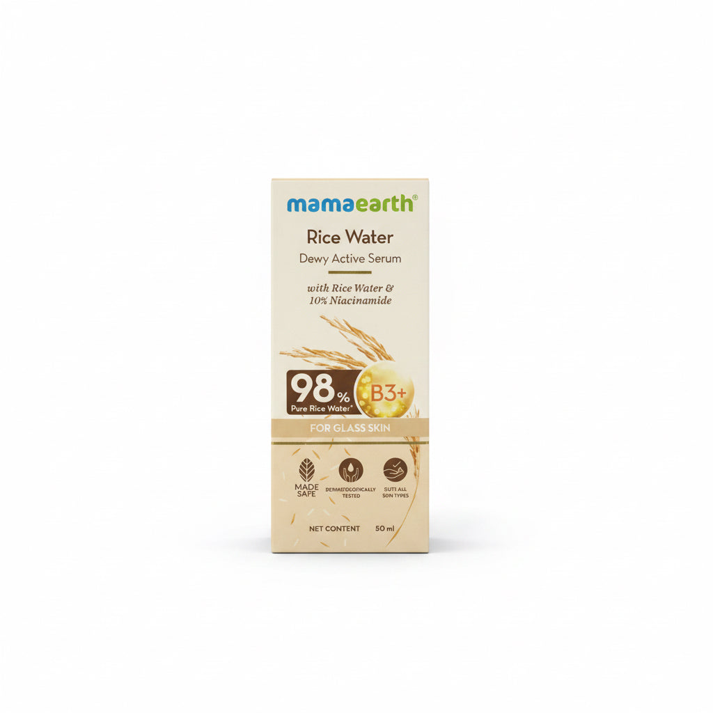 A box of Mama Earth Rice Water Dewy Active Serum stands on a red desk in front of an open laptop. The packaging is beige with brown and green text and graphics, and the background shows a workspace setting with shelves and a computer.