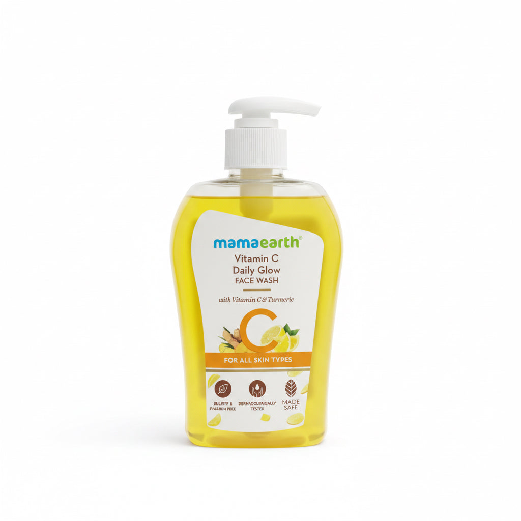 A bottle of Mama Earth Vitamin C Daily Glow Face Wash with a white pump dispenser, filled with yellow liquid, placed on a black counter next to a green basket containing other skincare products.