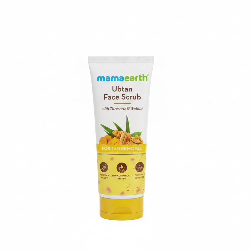 A tube of Mama Earth Ubtan Face Scrub with Turmeric & Walnut, placed on a counter next to a green basket and an open laptop in what appears to be a retail or office setting.