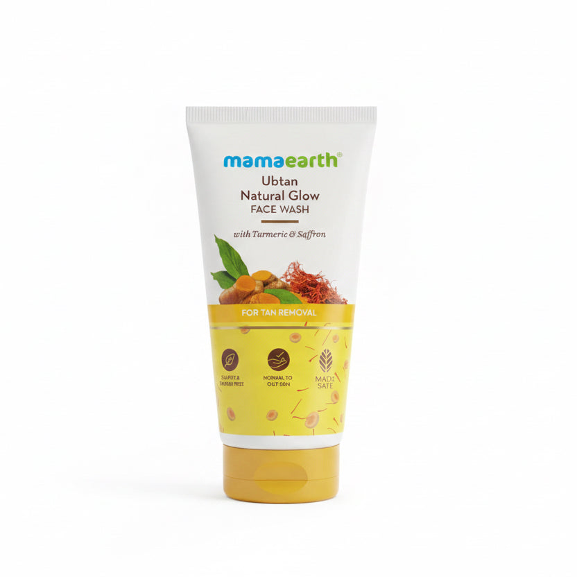 A tube of Mamaearth Ubtan Natural Glow Face Wash standing on a laptop keyboard in a workspace setting. The product packaging is cream and yellow with orange and red botanical illustrations, and the Mamaearth brand name is clearly visible.