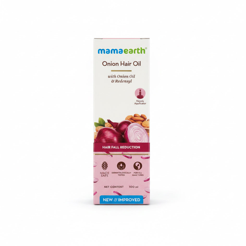A box of Mamaearth Onion Hair Oil standing on a reflective dark tabletop in an office-like setting. The packaging is mostly white with red and pink imagery of onions, almonds, and a hair icon, plus the Mamaearth brand name at the top.