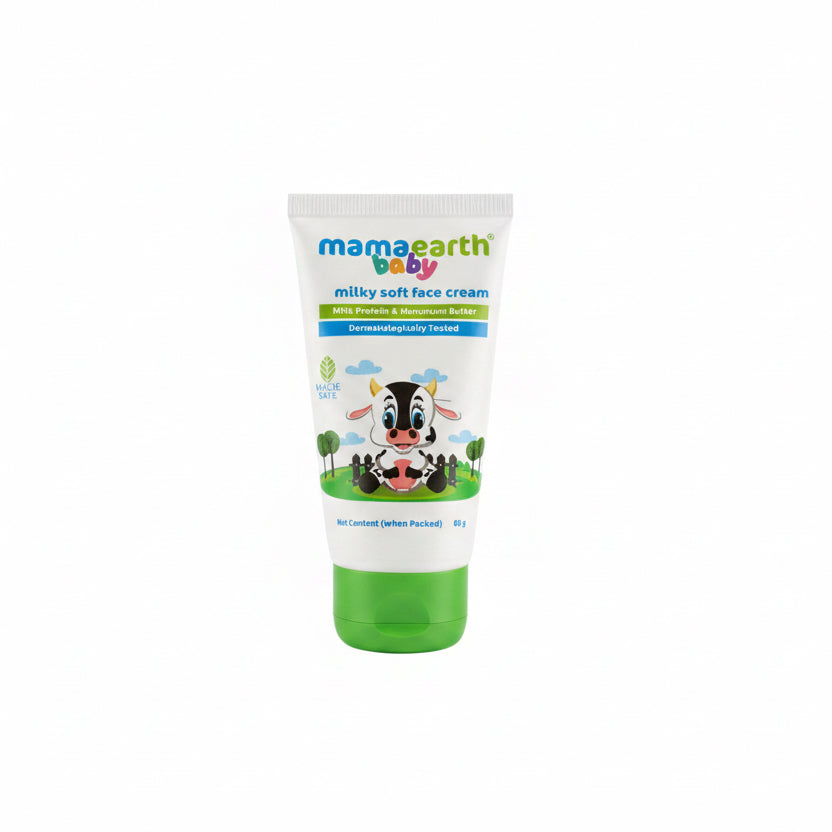 A tube of Mama Earth Baby Milky Soft Face Cream standing on a desk in front of an open laptop. The white tube features a green cap and a cartoon cow illustration with the brand name 'mamaearth baby' in colorful text.