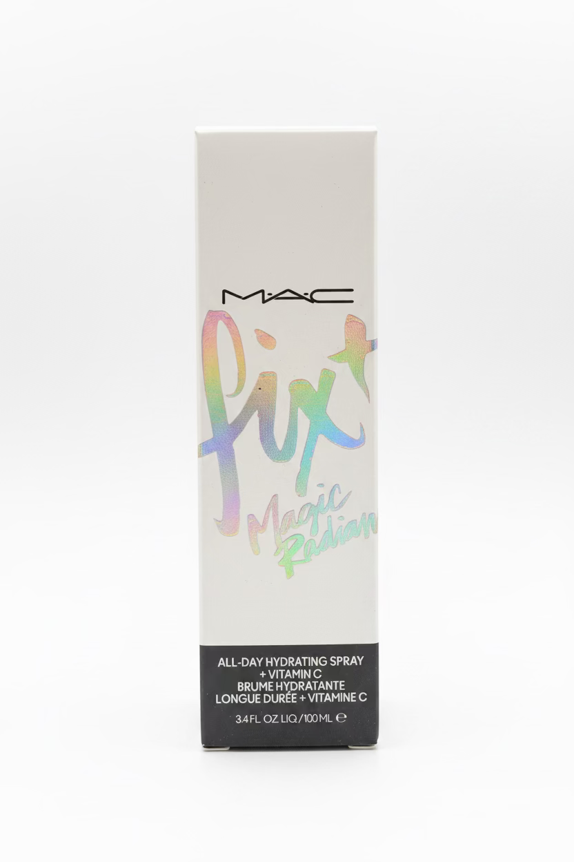 A box of MAC Fix Magic Radian All-Day Hydrating Spray standing on a glass display counter in a retail store. The packaging is white with a holographic pastel 'fix' script and a black text block at the bottom.