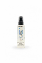 A bottle of MAC All-Day Magic Potion Humidifying Spray standing on a glass shelf in a retail store setting, with a laptop and shelves of products blurred in the background.