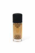 A bottle of MAC Studio Fix Fluid SPF 15 liquid foundation with a black cap, placed on a glass display counter in a retail store setting with a laptop and other products blurred in the background.