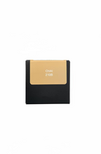 A hand holding a small square product box with a beige shade panel labeled 'CHAI 210B' against a blurred background of a person's legs and clothing.