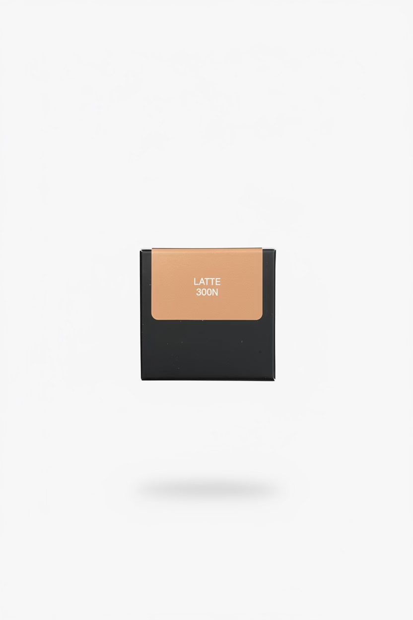 A hand holding a small square makeup compact or swatch box with a beige shade labeled 'LATTE 300N' on a black base, photographed over a person's lap with blue jeans and a plaid shirt visible.
