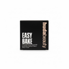 A black square box of hudabeauty Easy Bake loose setting powder, placed on a glass display counter in a retail store with other products blurred in the background.