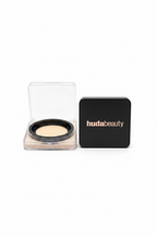 A close-up of a hudabeauty compact powder case next to an open square compact showing a cream-colored powder, displayed on a glass shelf in a retail setting.