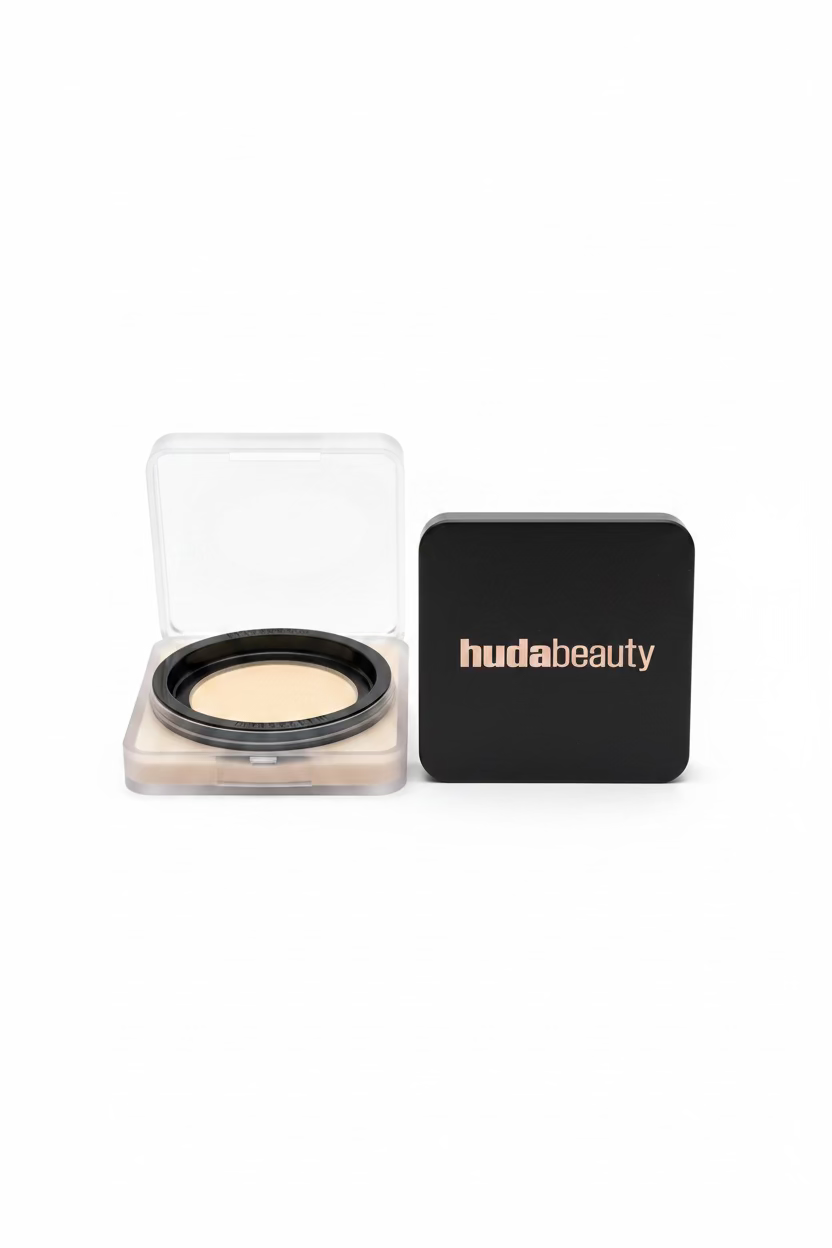 A close-up of a hudabeauty compact powder case next to an open square compact showing a cream-colored powder, displayed on a glass shelf in a retail setting.