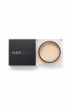 A hand holding a Huda Beauty pressed powder foundation compact and its black outer box, with the brand name 'HUDA BEAUTY' visible on the box.