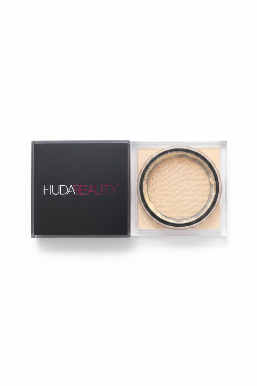 A hand holding a Huda Beauty pressed powder foundation compact and its black outer box, with the brand name 'HUDA BEAUTY' visible on the box.