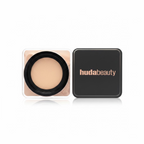 A Huda Beauty pressed powder compact shown open next to its black square case on a glass display shelf, with other cosmetics and retail items blurred in the background.