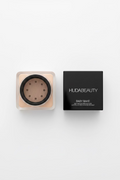 A square black and beige makeup compact or container set on a glass shelf, with the front piece showing a circular black center and gold dots, next to a black box labeled hudabeauty and BABY RAINE.